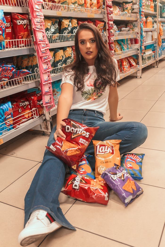About Young woman with trendy style sits on supermarket floor surrounded by snack bags.