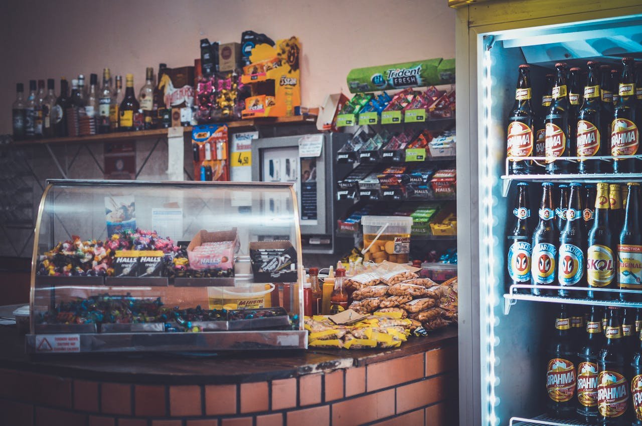 About A well-stocked convenience store counter with snacks, drinks, and a refrigerator showcasing beverages.